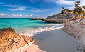 Playa del Carmen con rocas en primer plano, orilla de arena fina y vegetación tropical sobre un pequeño acantilado frente al mar