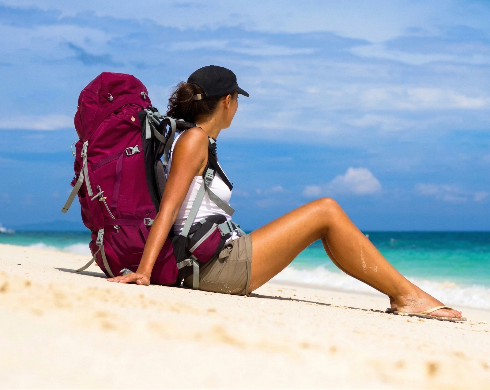 Joven mochilera viajando con una mochila de viaje color vino sentada sobre la arena blanca de una playa en la Riviera Maya, mirando hacia el mar turquesa.