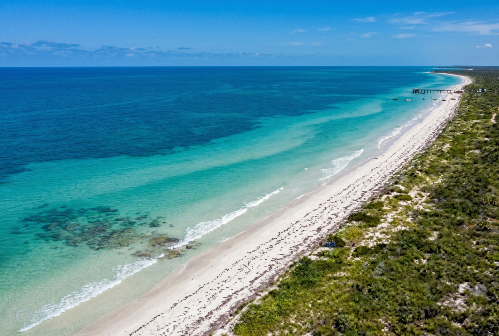 Vista aérea de la costa del Pueblo Mágico de Sisal en Yucatán, muestra el mar turquesa, arena blanca, arboles y un muelle en la playa.